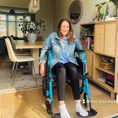 The image shows Charlotte Croft sitting in a blue wheelchair inside a well-lit, modern living room. She has long brown hair and is smiling. She is wearing a denim jacket over a blue top and black leggings, along with white socks. The room features a dining table with white chairs, a sideboard with decorative items, and a large round mirror on the wall. There is also a bookshelf with books and a vase of flowers. The Instagram handle @fnd.and.me8 is visible on the image. The overall atmosphere of the room is warm and inviting.