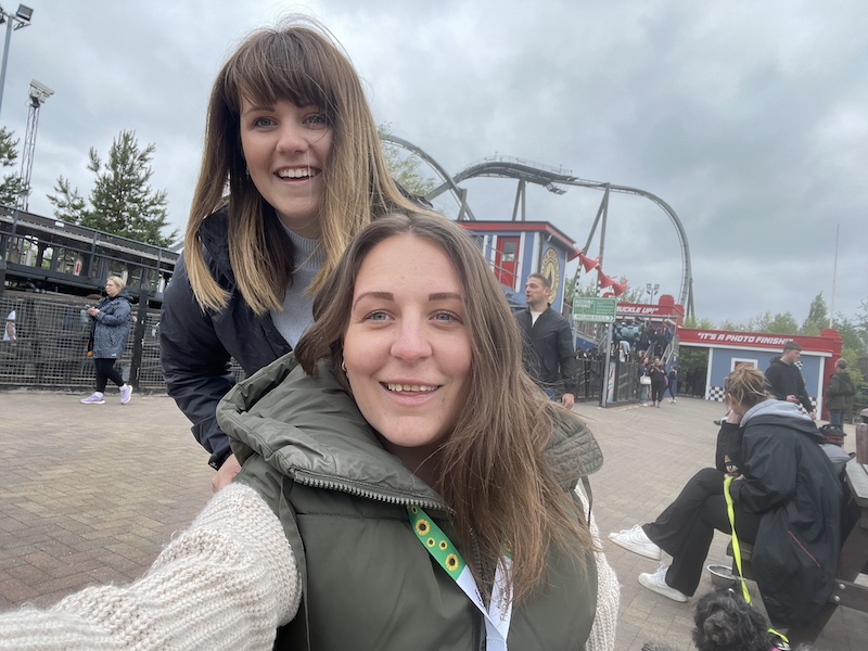 The photo features Charlotte sat in her wheelchair with her partner standing behind her, both smiling at the camera. Charlotte is wearing a green jacket and a cream-coloured sweater. The woman behind her (Charlotte’s partner) is wearing a black jacket and has shoulder-length brown hair with bangs. They appear to be at an amusement park with a roller coaster visible in the background. Other people can be seen in the background as well, including a woman walking and a man standing near the roller coaster entrance. The sky is overcast, suggesting a cloudy day.