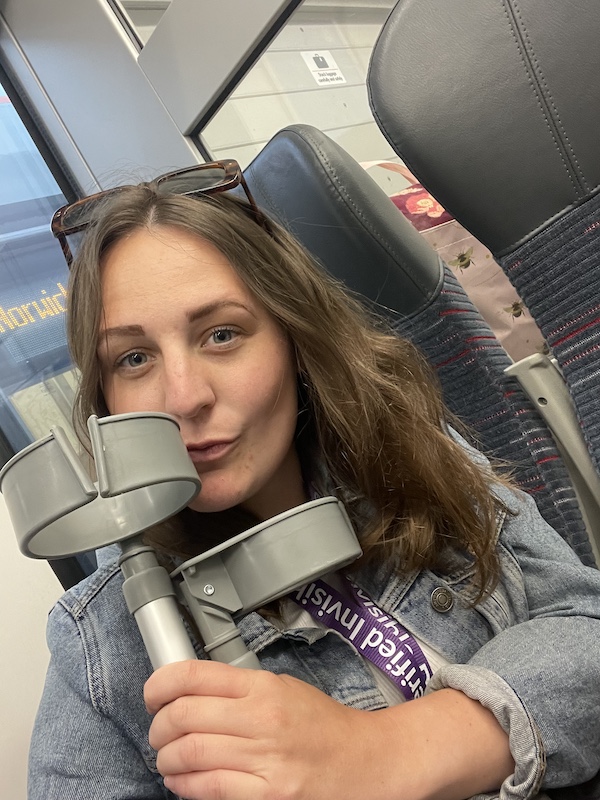 The image shows Charlotte with long brown hair, wearing a denim jacket and sunglasses on her head, sitting in a train seat. She is holding a pair of gray crutches close to her face and making a playful kissy face. She has a lanyard around her neck with text on it that appears to say "Certified Invigilator." The train interior is visible in the background, including the window and seat upholstery.