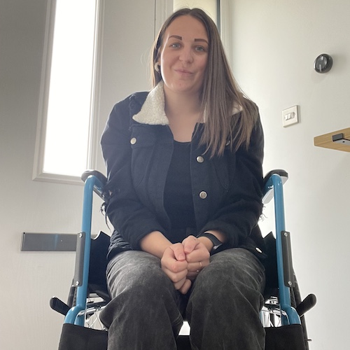 Charlotte in a wheelchair. She has long brown hair and is wearing a black jacket with a white sherpa collar, a black top, and dark pants. She has her hands clasped together on her lap and is smiling slightly at the camera. The background appears to be the entrance of a house, with a door and window visible behind her, letting in natural light. There is also a thermostat or some type of control on the wall to her right.