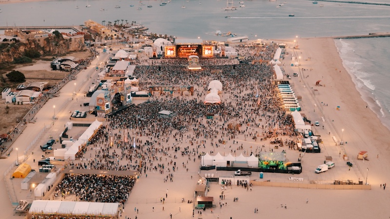 The image shows a large beach music festival, likely taken from a drone. A massive crowd is gathered around a brightly lit stage near the top center. The festival area is filled with tents and stalls, organized in rows. The beach and ocean are visible on the right, with boats anchored offshore. The warm lighting suggests the photo was taken around sunset.