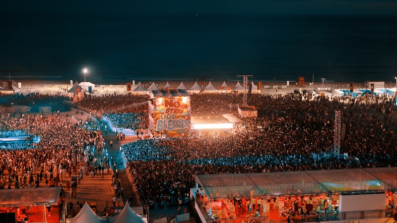 The image shows a nighttime beach music festival, with a large crowd gathered in front of a stage that displays a colorful banner reading "No Stress Just Bless." The stage is located near the center of the image, with bright lights illuminating the area. The ocean is visible in the background, and tents line the back of the festival grounds. The crowd is densely packed, and the scene is vibrant with various lighting effects, giving the event an energetic atmosphere.