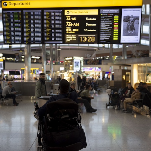 The image depicts an airport departure lounge. A large yellow and black departure board dominates the top portion, showing various flight details such as gate numbers and departure times. Below the board, the terminal is bustling with travelers, some seated and others walking around. In the foreground, a person in a wheelchair is facing the departure board, holding a smartphone. The overall atmosphere is busy, with numerous passengers and airport staff visible in the background, highlighting the typical activity and movement in an airport setting.