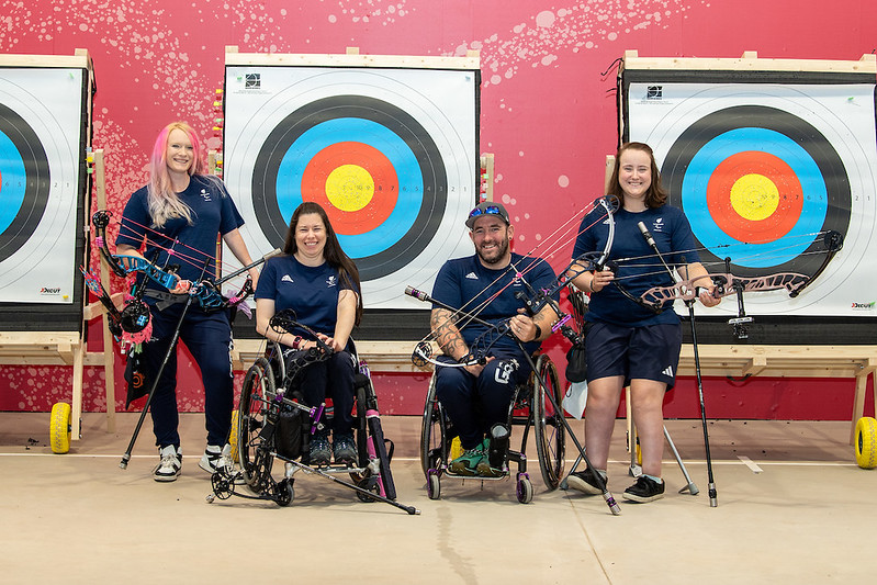 The image features four members of the ParalympicsGB Para Archery team posing in front of two archery targets. The backdrop is a vibrant red wall with splatter designs. - **Left to Right**: 1. **First Archer**: Jodie Grinham A woman with long, light pink hair stands on the far left. She is holding a bow and smiling, wearing a navy blue shirt and pants. 2. **Second Archer**: Victoria Kingstone, seated in a wheelchair, is in the middle left position. She is also holding a bow and wearing a navy blue shirt and pants, smiling warmly. 3. **Third Archer**: Nathan MacQueen is seated in a wheelchair next to Victoria. He is wearing a navy blue cap, shirt, and pants, holding his bow and smiling broadly. 4. **Fourth Archer**: Phoebe Paterson Pine stands on the far right, holding a bow and smiling. She is dressed similarly in a navy blue shirt and shorts. All archers are wearing similar navy blue outfits with the ParalympicsGB branding. The archery targets behind them feature standard colored rings, and all appear to be in a training or competition setting.