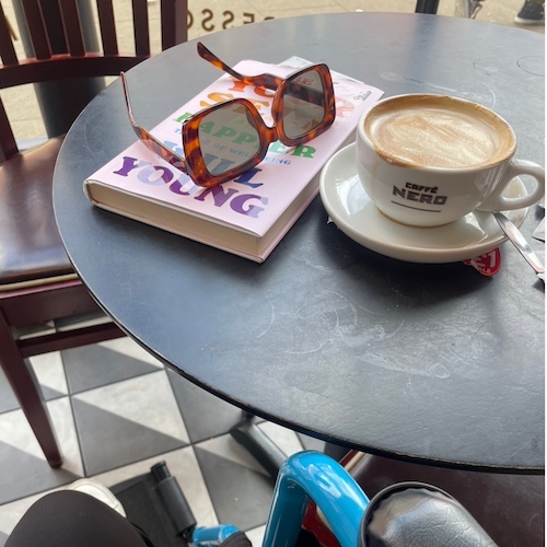 The image shows a round café table at Caffè Nero. On the table, there is a book with a colorful cover featuring large, bold text. A pair of tortoiseshell sunglasses is resting on top of the book. Next to the book, there is a cup of coffee with a frothy top, served in a white cup and saucer with the Caffè Nero logo printed on the cup. The table is black, with some faint chalk-like marks or scratches on the surface. Part of a checkered floor, a wooden chair, and the lower half of someone's leg can be seen in the background. The overall setting suggests a casual, relaxing moment in a café.