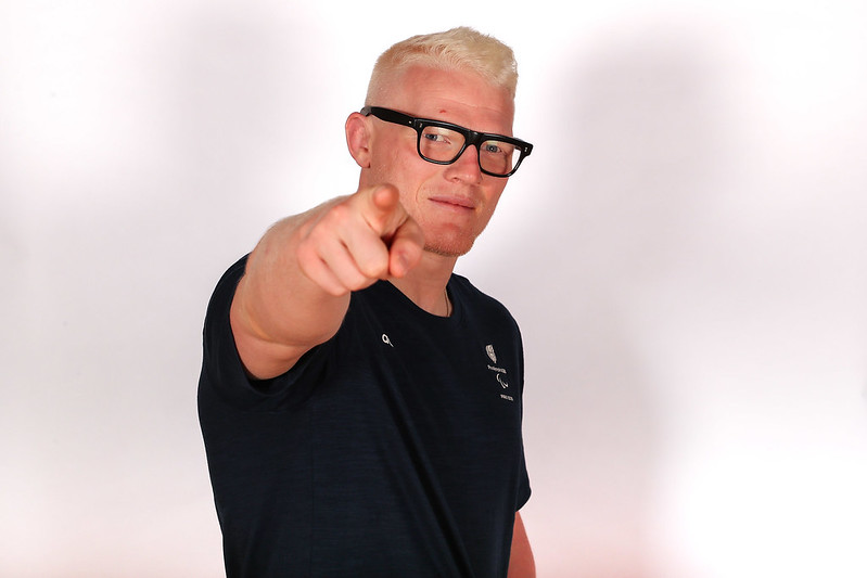 The image shows Evan Molloy standing against a plain, light-colored background. He has short, light blonde hair and is wearing black-framed glasses. He is smiling and wearing a navy blue Adidas T-shirt, which features the ParalympicdGB Paris 2024 logo on the left side of the chest. Evan has an athletic build and is standing with one arm behind his neck.