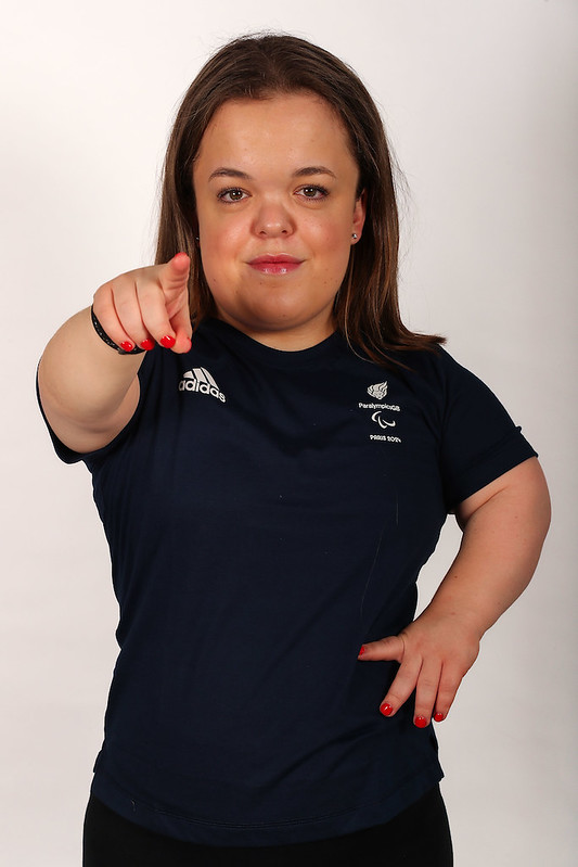 The image shows Lottie McGuiness with long brown hair, wearing a navy blue T-shirt with the logos of Adidas and ParalympicsGB printed on it. The ParalympicsGB logo includes the Agitos symbol, which is associated with the Paralympic Games. Lottie is smiling and pointing her right hand at the camera. The photo has a clean, light background, drawing attention to her face and the details on her shirt.