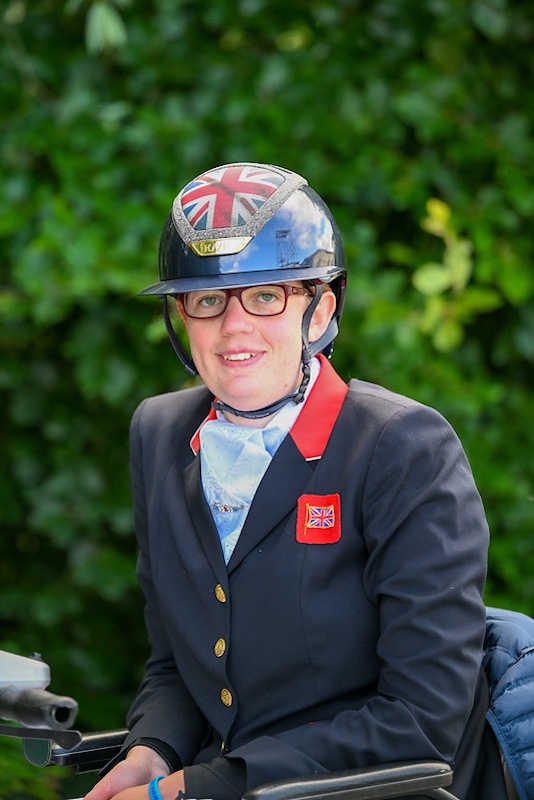 The image shows Mari Durward-Akhurst wearing equestrian attire, including a helmet and a formal jacket. The helmet has a Union Jack design on it. She is wearing glasses and has a light-colored stock tie under her jacket, which is adorned with red and white accents and features a badge with a Union Jack flag. The woman is seated in a wheelchair and is smiling at the camera. The background is a blurred outdoor setting with green foliage. The overall impression is of someone prepared for an equestrian event, representing Great Britain.