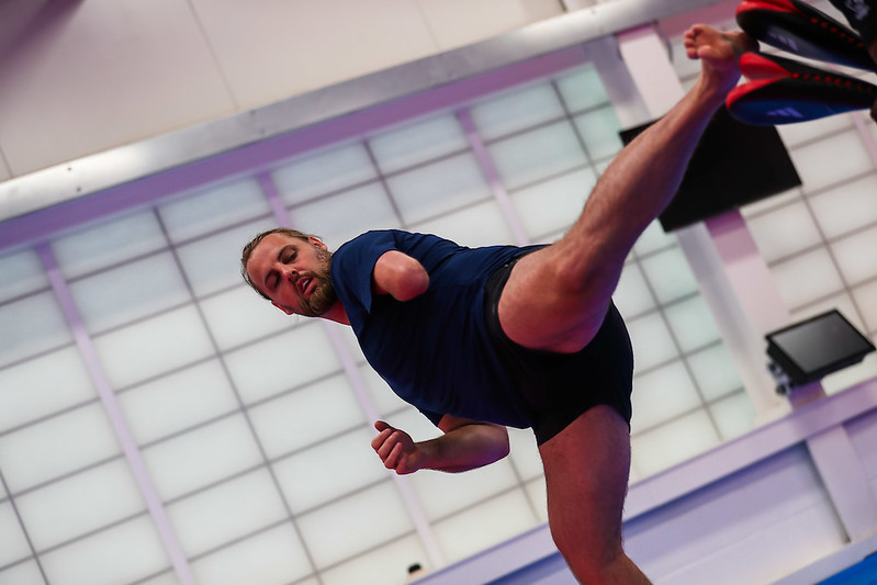 The image shows Matt Bush performing a high kick in a taekwondo training session. He is wearing a dark blue shirt and black shorts. The setting appears to be an indoor training facility with large, frosted windows in the background, which provide a diffused light. Matt's focus is on his kick, demonstrating balance and athleticism. The image captures a dynamic moment, highlighting his skill and dedication to the sport.