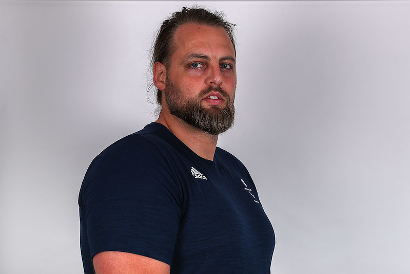 The image shows bearded Matt Bush looking sideways and smiling. He is wearing a dark blue Adidas t-shirt with the ParalympicsGB logo. The background of the image is plain and light-coloured.