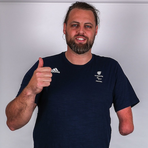 The image shows Matt Bush giving a thumbs-up gesture. He has a beard and is smiling. He is wearing a dark blue Adidas t-shirt with the ParalympicsGB logo and Paris 2024. The background of the image is plain and light-coloured.