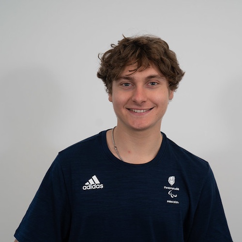 The image shows Matthew Robertson with short, slightly curly brown hair, smiling at the camera. He is wearing a navy blue Adidas t-shirt with logos on the chest, including the Paralympics symbol and text that appears to relate to a Paralympic event or team. The background is plain white, giving a professional and clean look to the portrait. The individual appears friendly and approachable, with a casual and sporty style.