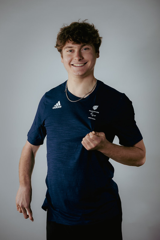 The image shows Matthew Robertson with short, slightly curly brown hair, smiling at the camera. He is wearing a navy blue Adidas t-shirt with logos on the chest, including the Paralympics symbol and text that appears to relate to a Paralympic event or team. The background is plain white, giving a professional and clean look to the portrait. The individual appears friendly and approachable, with a casual and sporty style.