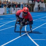 The image shows an athlete participating in a wheelchair race on a bright blue track. The athlete, dressed in a red shirt with black sleeves and a red helmet, is focused and determined as they push their wheelchair forward. The wheelchair is specially designed for racing, with a streamlined structure and a low-to-the-ground profile, equipped with two large rear wheels and a smaller front wheel. The athlete is leaning forward, propelling the wheelchair with their gloved hands. In the background, a crowd of spectators can be seen, watching the race intently. Some of them are slightly blurred due to the focus on the athlete, emphasizing the movement and speed of the scene. The track lines are clearly visible, curving slightly, indicating that the athlete is navigating a bend. The atmosphere appears to be competitive and energetic, highlighting the intensity of the race. The overall color palette is vibrant, with the blue track contrasting sharply against the athlete's red attire.