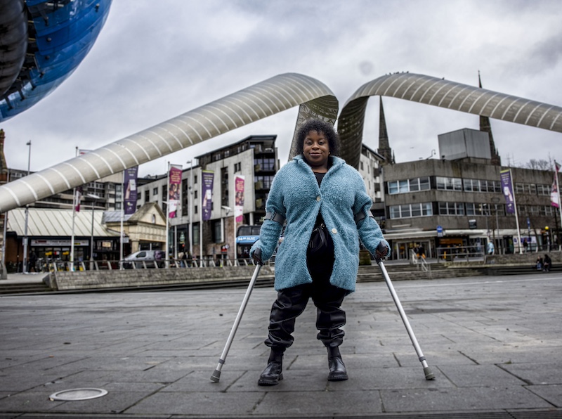 The image shows a woman standing confidently with the aid of two crutches in a public square. She is wearing a light blue coat and black pants, and has a warm smile. The background features a distinctive architectural structure with two large, curved, metallic arcs that frame the scene. There are several modern buildings, banners, and some people in the vicinity. The sky is overcast, suggesting it is a cloudy day. The overall setting appears to be urban and contemporary.