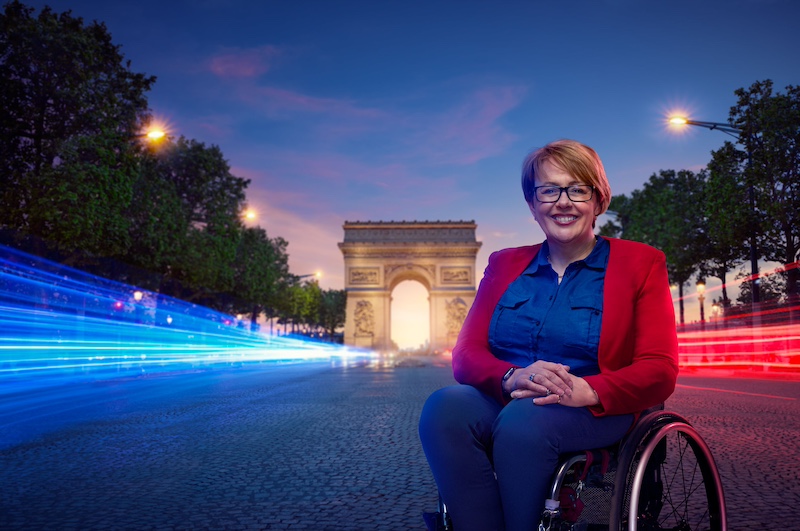 The image features Tanni Grey-Thompson in her wheelchair positioned prominently in the foreground. She has short blonde hair, glasses, and a friendly smile. She is wearing a blue shirt with a red blazer over it. The background shows the Arc de Triomphe, a famous monument in Paris, France, with a street illuminated by dynamic light trails that suggest motion, likely from passing vehicles, adding a sense of energy and movement to the scene. The lighting suggests that this is taken during the evening or twilight, with the sky displaying a deep blue hue.