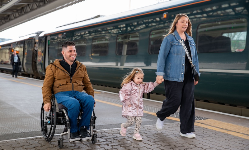 The image captures a family at a train station platform. A man in a wheelchair, wearing a brown jacket and blue jeans, is smiling and moving along the platform. Next to him, a woman in a denim jacket and black pants holds the hand of a young girl in a pink coat and patterned leggings. The young girl, also smiling, is walking between the man and the woman. In the background, a modern train is stationed at the platform, and the scene appears to be set during the daytime. The family seems to be enjoying their time together, and the atmosphere is cheerful and relaxed.