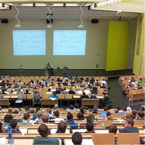 The image shows a large university lecture hall filled with students attending a lecture. The seating is arranged in rows with long desks, and most seats are occupied by students who are focused on the lecture. The lecturer, positioned at the front center, is standing behind a podium and is likely explaining the content displayed on two large projection screens behind them. The screens show complex mathematical equations or formulas, suggesting that the lecture is likely on a technical or scientific subject, possibly mathematics or physics. The hall has a modern design with high ceilings, and the walls are light-colored with yellow panels on either side of the screens. The students appear engaged, with some taking notes, using laptops, or looking at the screen. The atmosphere seems to be one of concentrated learning, typical of a university setting.