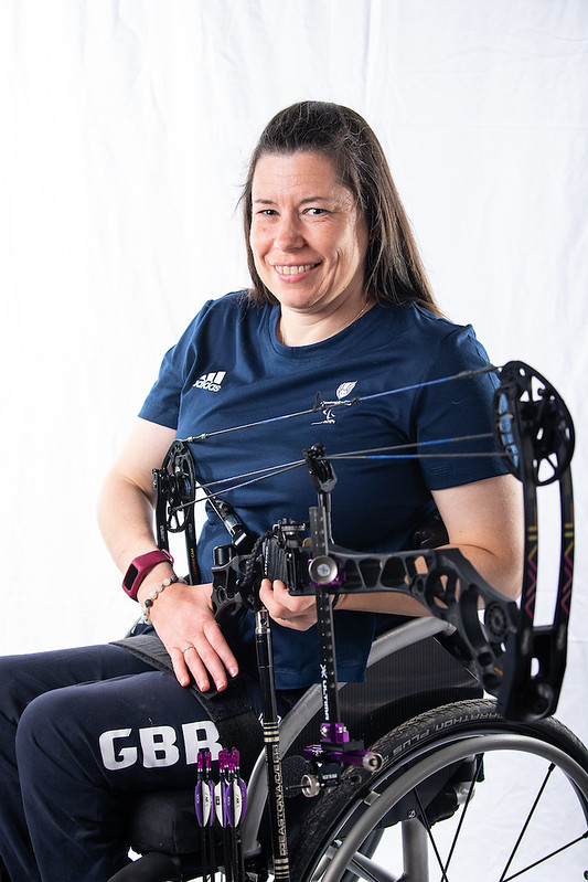 The image shows Victoria Kingstone seated in a wheelchair. She has long brown hair and is smiling warmly. She is wearing a navy blue shirt with ParalympicsGB Paris 2024 logo on the chest. Her trousers are also navy blue and feature the letters "GBR" prominently. The background of the image is plain white, drawing full attention to the subject.