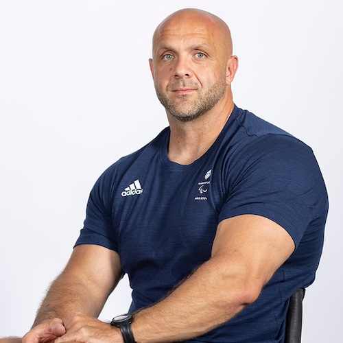 The image shows Ed Clifton, a strong, muscular man sitting in a wheelchair. He is bald, with a neatly trimmed beard and a focused expression on his face. He is wearing a navy blue Adidas T-shirt that appears to have a Paralympics logo, His arms are crossed in front of him, displaying his strong build, and he wears a black watch on his left wrist.