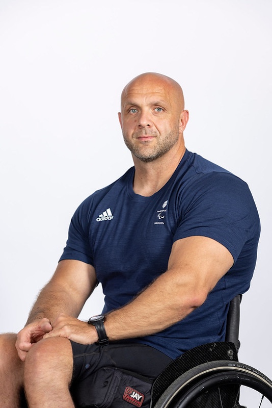 The image shows Ed Clifton, a strong, muscular man sitting in a wheelchair. He is bald, with a neatly trimmed beard and a focused expression on his face. He is wearing a navy blue Adidas T-shirt that appears to have a Paralympics logo. His arms are crossed in front of him, displaying his strong build, and he wears a black watch on his left wrist. The wheelchair is visible at the bottom of the image, and he is seated in a studio setting with a plain white background.