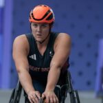 The image shows Eden Rainbow Cooper, a female athlete in a wheelchair, wearing a bright orange helmet and a dark athletic uniform that reads "Great Britain." She appears to be focused, with her hands in front of her, likely preparing for a racing event. Her expression is determined and concentrated, emphasising her competitive spirit.