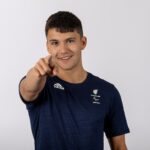 The image shows Henry Urand, a young man with short dark hair, smiling and pointing directly at the camera. He is wearing a navy blue Adidas t-shirt with the logo of "Paralympics GB" and another smaller logo that appears to be related to the Paralympic Games. The background is plain and white, emphasizing the subject. Henry appears to be friendly and engaging, with an athletic build and a positive demeanor.