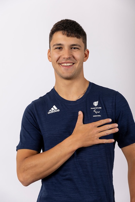 The image shows Henry Urand, a young man with short dark hair, smiling and pointing directly at the camera. He is wearing a navy blue Adidas t-shirt with the logo of "Paralympics GB" and another smaller logo that appears to be related to the Paralympic Games. The background is plain and white, emphasizing the subject. Henry appears to be friendly and engaging, with an athletic build and a positive demeanour.