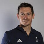 The image shows Jaco van Gass with short, dark hair, a light complexion, and a friendly expression. He is wearing a dark navy polo shirt with logos on it, including the Adidas logo and another logo featuring a fern and the word "Paralympics." The background is plain and neutral, likely a studio or professional setting, suggesting that the photo is part of a formal portrait or a promotional image.