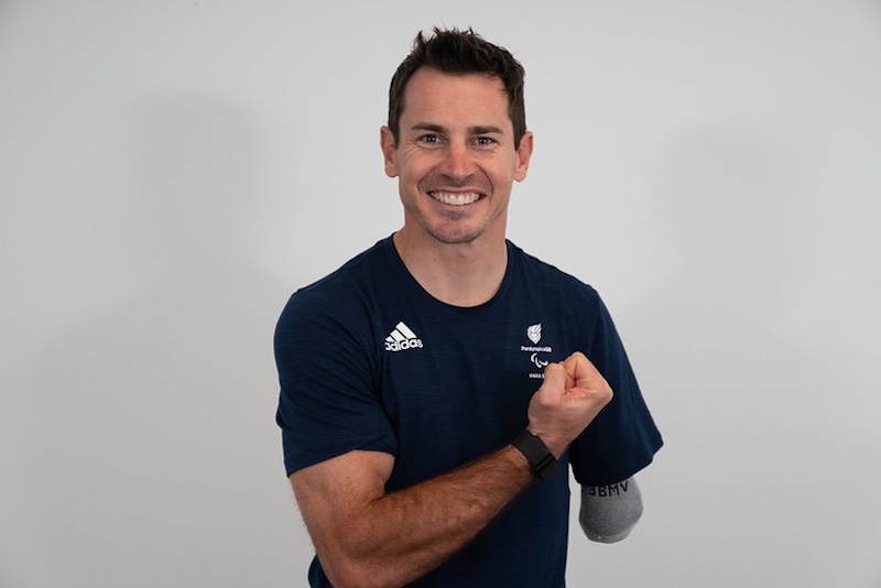 The image shows Jaco van Gass with short, dark hair, a light complexion, and a friendly expression. He is wearing a dark navy polo shirt with logos on it, including the Adidas logo and another logo featuring a fern and the word "Paralympics." The background is plain and neutral, likely a studio or professional setting, suggesting that the photo is part of a formal portrait or a promotional image.