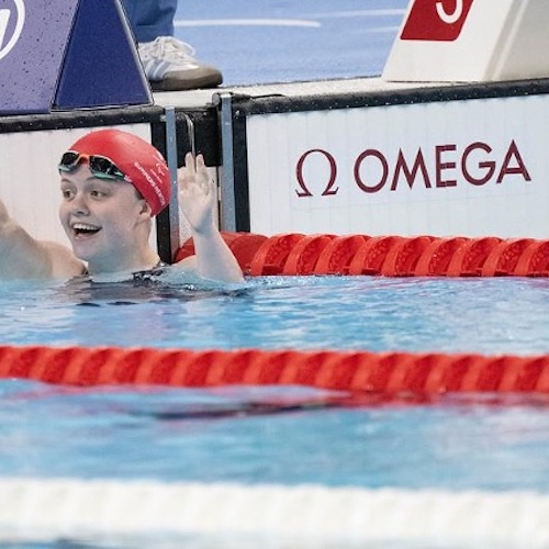 The image shows Maisie Summers-Newton, a swimmer wearing a red swim cap and goggles, smiling and raising a hand in a gesture of celebration or greeting. Maisie is in a swimming pool, holding onto the edge of the pool near a wall that has the logo of "OMEGA," a well-known sports timekeeping company. The water is clear, and there are red lane dividers in the pool. The atmosphere suggests that Maisie has just finished a race and is pleased with the outcome.