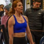 The image shows Rachel Hives engaged in a fitness activity, possibly in a gym environment. She has red hair and is wearing a blue sports bra and black athletic leggings or shorts. Her posture and facial expression suggest she is focused, likely in the midst of a workout or exercise session. The background includes other people, some of whom are wearing warm clothing such as jackets and beanies, implying the gym might be in a cooler climate or that people are waiting for their turn. The scene appears to be a CrossFit or high-intensity workout session, based on the atmosphere and the athletåic wear.