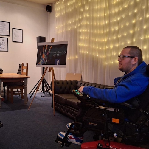 The image shows DK in a powered wheelchair wearing a blue jacket, glasses, and sneakers with red, white, and blue coloring. They appear to be engaged in a conversation or a presentation, facing someone out of view to the left. Behind them, there is a couch and a background decorated with string lights, creating a warm ambiance. To the side, an easel holds a poster with "Accessibility Arts" written on it, accompanied by an image of hands holding paintbrushes, suggesting a setting related to art, creativity, or inclusivity. The room is decorated simply, with framed prints on the wall.