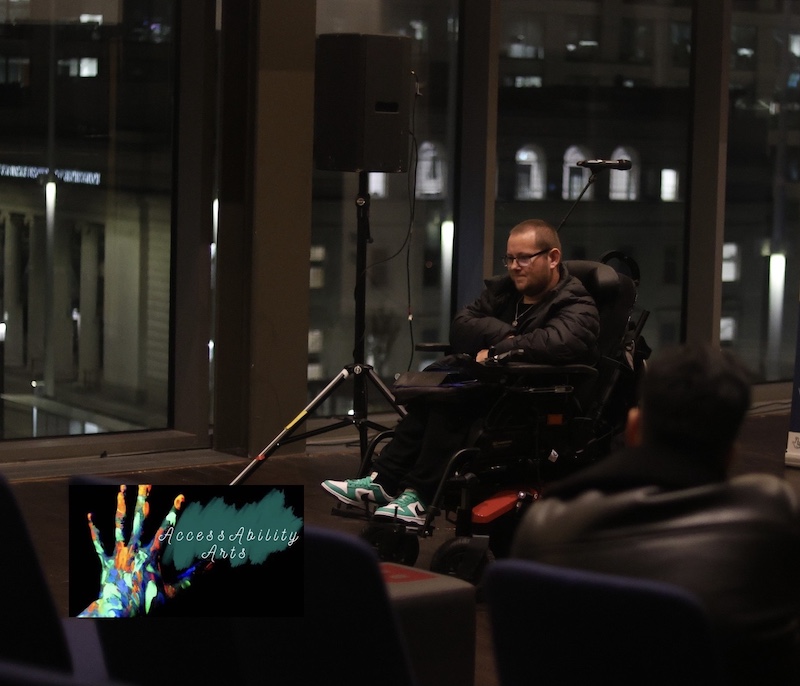 The image shows DK in a powered wheelchair speaking or presenting at what seems to be an indoor event. He is wearing glasses, a dark jacket, and green and white sneakers. The setting appears to be a modern venue, with large windows revealing an urban night scene outside, including illuminated buildings in the background. In the lower left corner of the image, there is a graphic overlay that shows a colorful handprint with the words "AccessAbility Arts" written in a stylized font. This suggests the event may be related to accessibility, inclusion, or the arts, possibly focused on raising awareness or providing a platform for artists with disabilities. In the foreground, there are blurred figures seated, suggesting an audience watching the speaker.