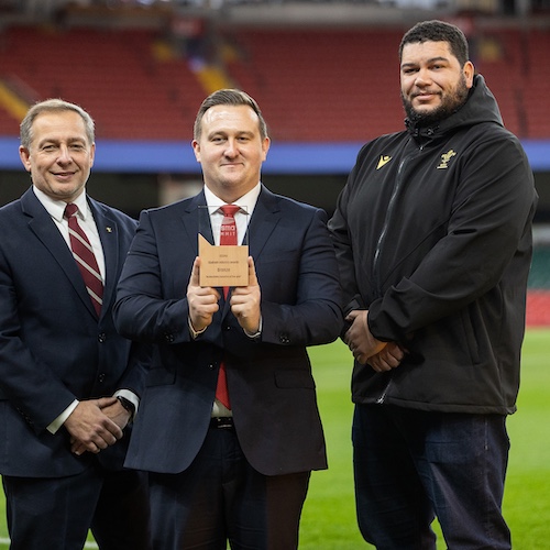 The image shows three individuals standing on a stadium field. The central figure, holding an award plaque, is flanked by two others. They appear to be in a formal setting, possibly celebrating an achievement related to the Principality Stadium's recognition for accessibility innovation. The background shows part of the stadium seating, adding to the sense of the location being a sports venue.