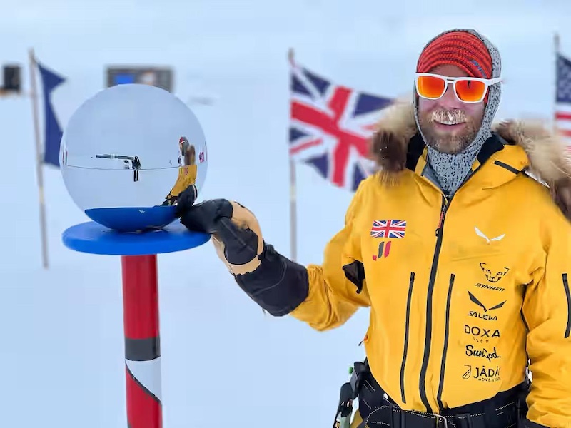 The image depicts Jonny standing next to the ceremonial marker for the Geographic South Pole in Antarctica. He is wearing a bright yellow parka with fur-lined hood, featuring multiple logos and patches, including a Union Jack. He is also dressed in cold-weather gear such as gloves, goggles with orange-tinted lenses, and a warm head covering. He is smiling and pointing or touching a reflective metallic sphere mounted on a red and white striped pole, which is the ceremonial marker for the South Pole. Flags of various countries, including the United Kingdom, are visible in the background, symbolizing international presence at the site. The environment is completely covered in snow, with clear skies.