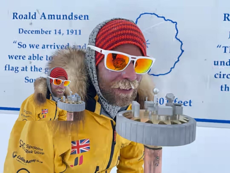 A close-up of Jonny stood by the South Pole monument. He is dressed in heavy polar gear, including a bright yellow parka with fur trim on the hood, clearly prepared for extreme cold weather conditions. He is also wearing protective goggles with a red tint. 