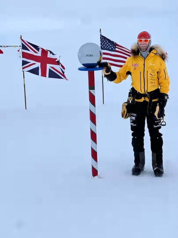 The image shows Jonny standing next to the ceremonial marker for the Geographic South Pole. He is dressed in cold-weather gear, including a bright yellow parka with a fur-lined hood, black insulated pants with sponsor logos, and gloves. They are also wearing red-tinted goggles and a striped head covering. He is holding or touching a reflective metallic sphere mounted on a red and white striped pole, which symbolizes the ceremonial South Pole. Behind him are flags of various nations, including the Union Jack (United Kingdom) and the United States flag, representing international cooperation. The entire scene is set in a snowy, icy environment under clear skies, typical of Antarctica.