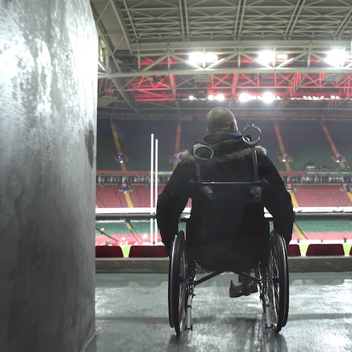 The image shows a person in a wheelchair overlooking a large sports stadium, likely from an accessible seating area. The stadium features prominent goalposts and brightly lit seating, with a roof structure illuminated by red lights. The individual is seen from behind, with a focus on their silhouette, emphasizing accessibility and inclusion in public spaces like stadiums. The atmosphere appears calm, suggesting the scene is either before or after an event.