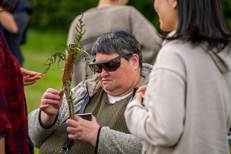 The image shows a thoughtful moment during an outdoor activity session. At the center, a person wearing dark wraparound sunglasses and a cozy, textured green sweater under a grey fleece jacket is carefully examining a handmade bundle crafted from natural materials — a stick adorned with green ferns and a large brown leaf, likely tied together.The individual holds a small paper cup in one hand, possibly used for collecting or supporting the materials, while engaging with others around them. Two other people are partly visible in the frame, one on each side, suggesting a collaborative or interactive atmosphere. The background shows lush greenery, indicating the activity is taking place in a natural setting like a garden, park, or research field. The person’s expression appears focused and reflective, highlighting their active participation and sensory engagement with the project.