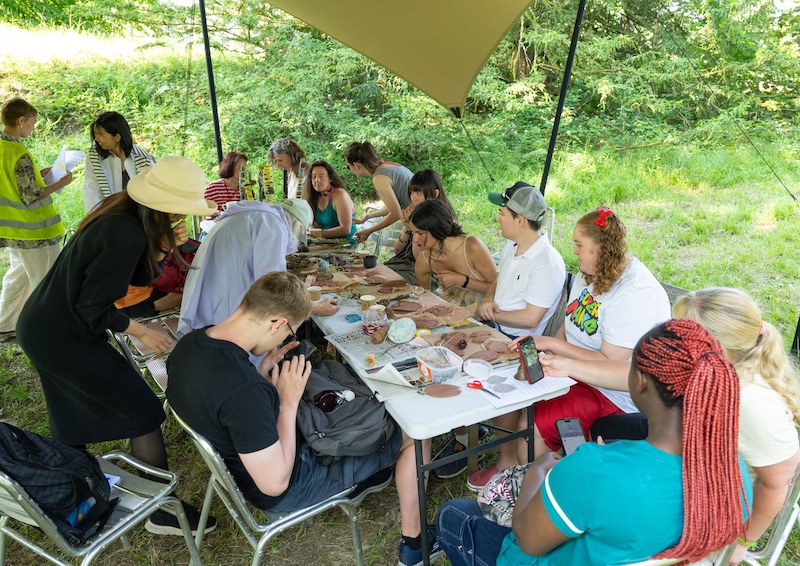 A group of people are gathered under a canopy tent in a grassy outdoor setting, participating in an arts and crafts workshop. They are seated around a long table covered with clay, sculpting tools, and art materials. Participants of various ages and backgrounds are engaged in creating clay pieces, some deeply focused while others chat or check their phones. A few facilitators stand nearby, offering guidance. The atmosphere is relaxed and collaborative, surrounded by greenery and dappled sunlight.