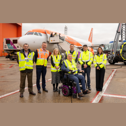 A group of seven individuals wearing high-visibility jackets stand on the tarmac in front of an easyJet aircraft at London Luton Airport. One individual is seated in a powered wheelchair at the centre of the group. The atmosphere is professional and welcoming, and the photo appears to be taken during an accessibility-focused airport event. Various airport vehicles and infrastructure are visible in the background.