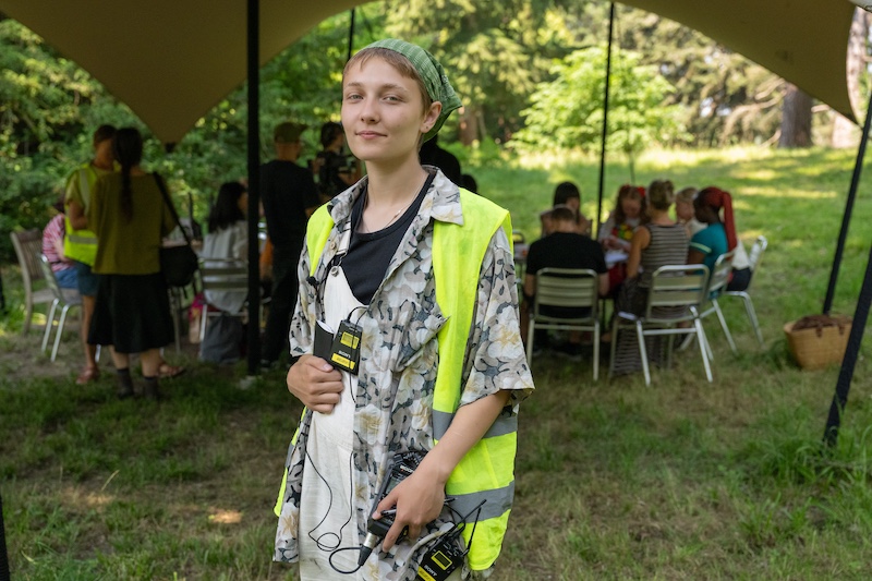 The individual - Maisie - has short hair and wears a green headscarf. They have a calm, confident expression, looking directly at the camera with a slight smile. In the background, several groups of people are seated around tables, actively engaged in conversations or activities. The setting is lush and green, with tall trees and grassy ground, giving the event a relaxed and nature-connected atmosphere.