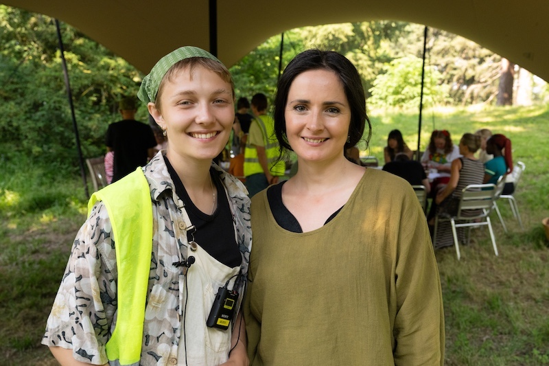 The photo shows two people standing side by side under a large outdoor canopy tent during a sunny day. Both individuals are smiling warmly at the camera, creating a friendly and welcoming atmosphere.On the left is a young person - Maisie - wearing a bright yellow high-visibility vest over a light patterned shirt. They also have a green headscarf and are carrying what looks like communication equipment — possibly a radio or microphone transmitter clipped onto their clothing. Standing next to them, on the right, is another individual - Karis - wearing a loose-fitting, earth-toned (olive green) top with a black undershirt. They have long, dark hair and a bright, engaging smile. In the background, under the same canopy, several people are gathered around tables, actively participating in what appears to be a workshop or community gathering. The setting is lush and natural, with green grass and tall trees surrounding the event space.