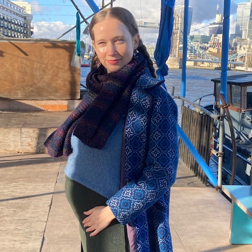 Victoria Bradley - A pregnant woman standing on a docked boat in London, smiling at the camera. She is wearing a patterned blue coat, blue sweater, and a dark scarf, with her hand gently resting on her baby bump. The Tower Bridge and river Thames are visible in the background under a sunny sky.