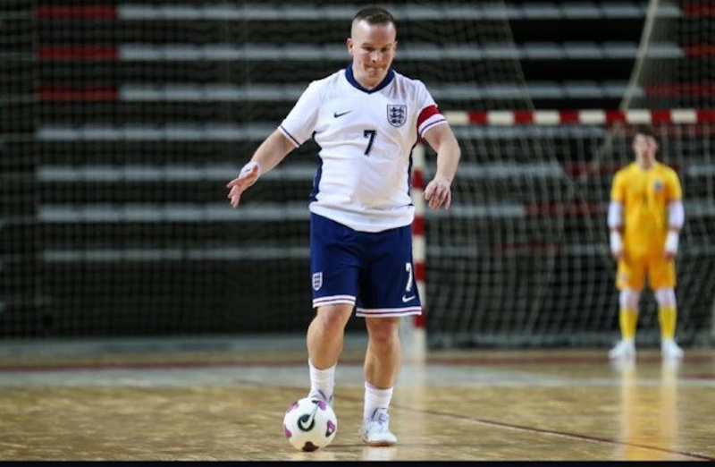 John McDougall wearing an England kit with jersey number 7 and a red captain's armband controls the ball on an indoor court. He is in motion, focused on the ball at his feet. Behind him, a goalkeeper in a yellow kit stands near the goal, which has a red and white net frame. The background shows empty bleachers.