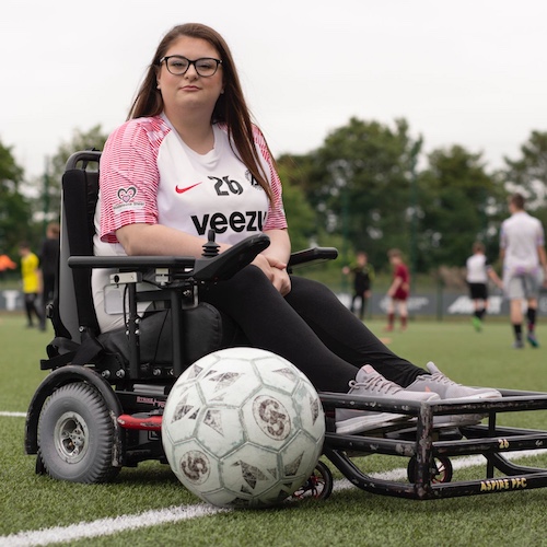 A young woman - Rosie Hodgson - in a powered wheelchair is on an outdoor football pitch, posing confidently with a large football positioned in front of her wheelchair’s foot guard. She is wearing a white jersey with red patterned sleeves, marked with the number 26 and the sponsor name “Veezu.” She has long brown hair, wears glasses, and has a composed expression. The wheelchair is equipped with a specialized front attachment for powerchair football. In the background, other players and spectators are visible on the pitch under an overcast sky, indicating an active game or event is taking place.