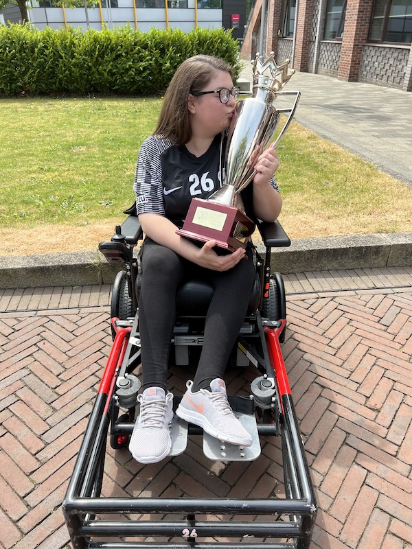 A young woman - Rosie Hodgson - in a power wheelchair is outdoors, kissing a large silver trophy with a crown-shaped top. She is wearing a black jersey with white-striped sleeves, number 26, and logos on the sleeves, including "Veezu." Her wheelchair is equipped with a red and black foot guard designed for powerchair football. She has long brown hair, glasses, and is dressed in black leggings and light grey Nike sneakers with orange swooshes. The background features a brick-paved path, a grass lawn, and a modern building with large windows. The moment captures a celebratory and triumphant mood.