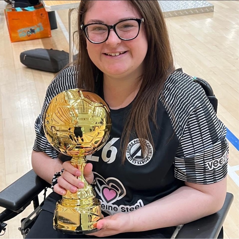 A smiling young woman in a wheelchair - Rosie Hodgson - holds a large golden trophy shaped like a football. She is wearing a black jersey with striped sleeves, the number 26, and several logos including "Aspire Powerchair Sports Club," "Veezu," and "Meine Steel." Her long brown hair is down, and she is wearing glasses. The setting appears to be an indoor sports hall with a light wooden floor. The trophy has a reflective surface and is engraved with text, signifying a personal or team achievement.