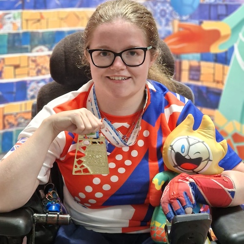 Claire Taggart, wearing a red, white, and blue Boccia UK jersey, smiles while holding up her 2022 World Championship gold medal and a colourful event mascot, seated in her wheelchair against a vibrant tiled backdrop.